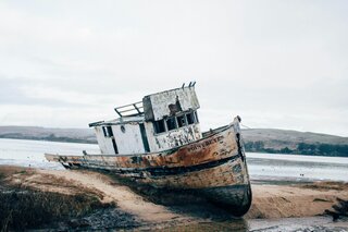 Ein altes, verfallenes Boot liegt auf dem Sand, teils im Wasser. Die umliegende Landschaft ist ruhig und leicht neblig.
